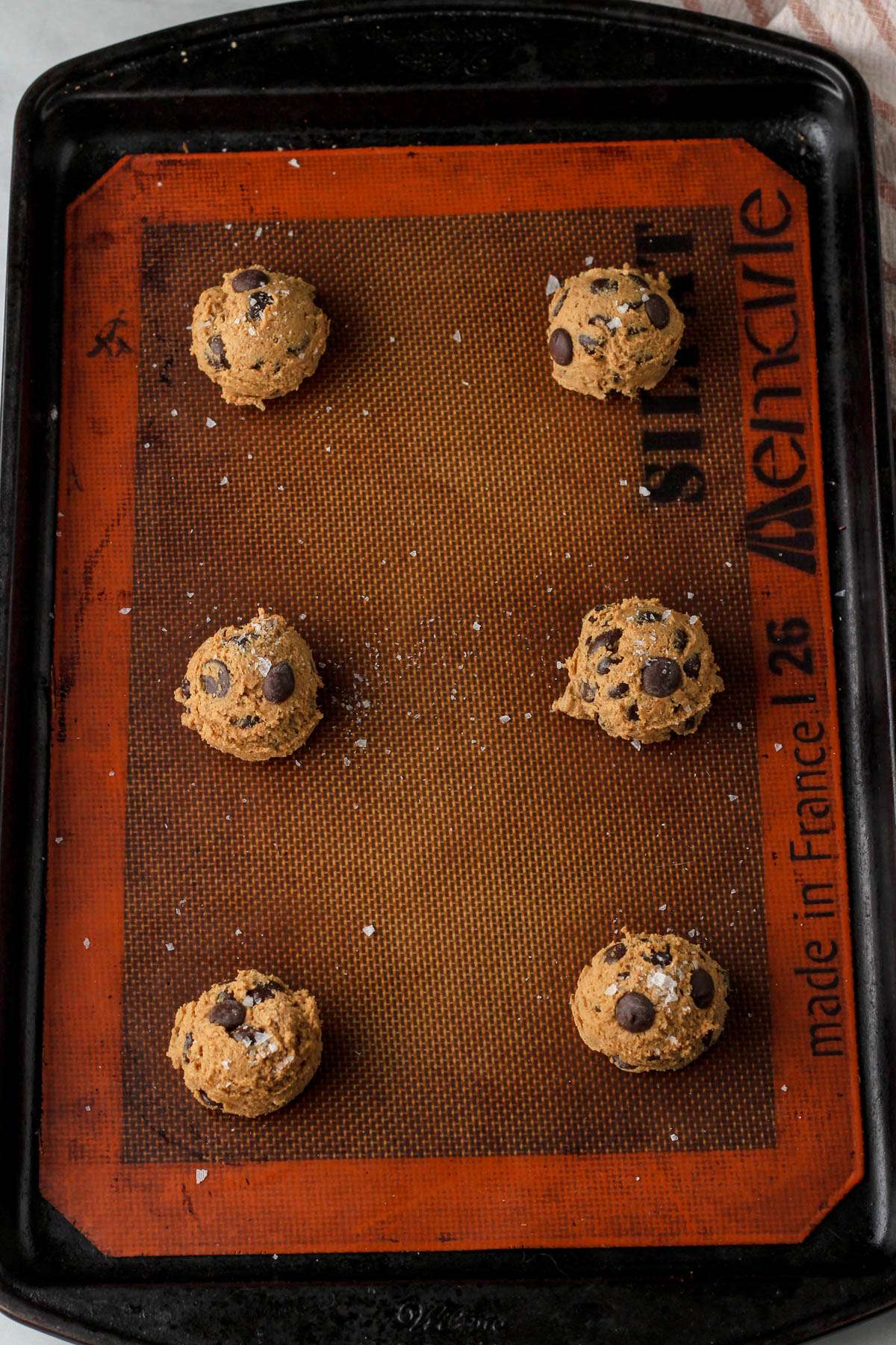 Espresso chocolate chip cookie dough balls topped with flaky sea salt on a silicone mat lined baking sheet before baking.