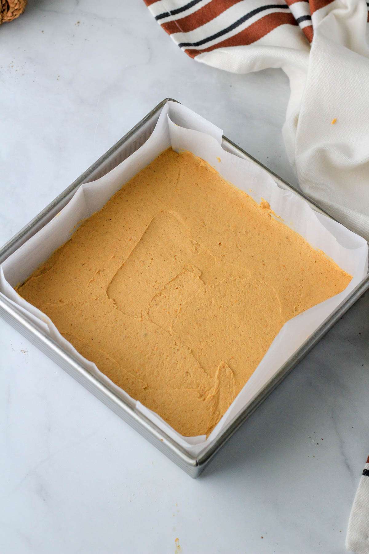 A silver square baking pan lined with parchment paper and the pumpkin spice cookie dough spread out smoothly before baking.