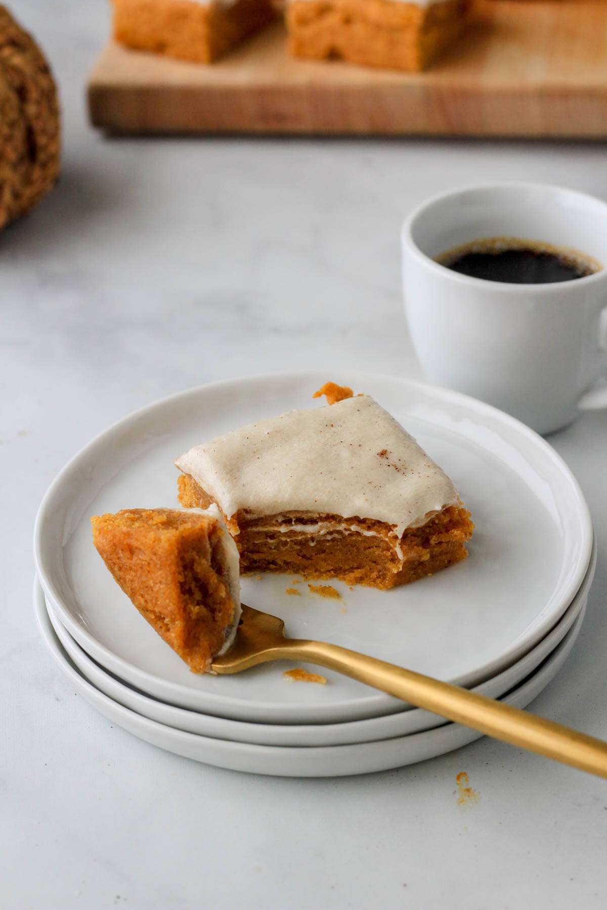A piece of frosted dairy-free pumpkin spice cookie bars on a stack of white plates with a small cup of coffee behind the plates.