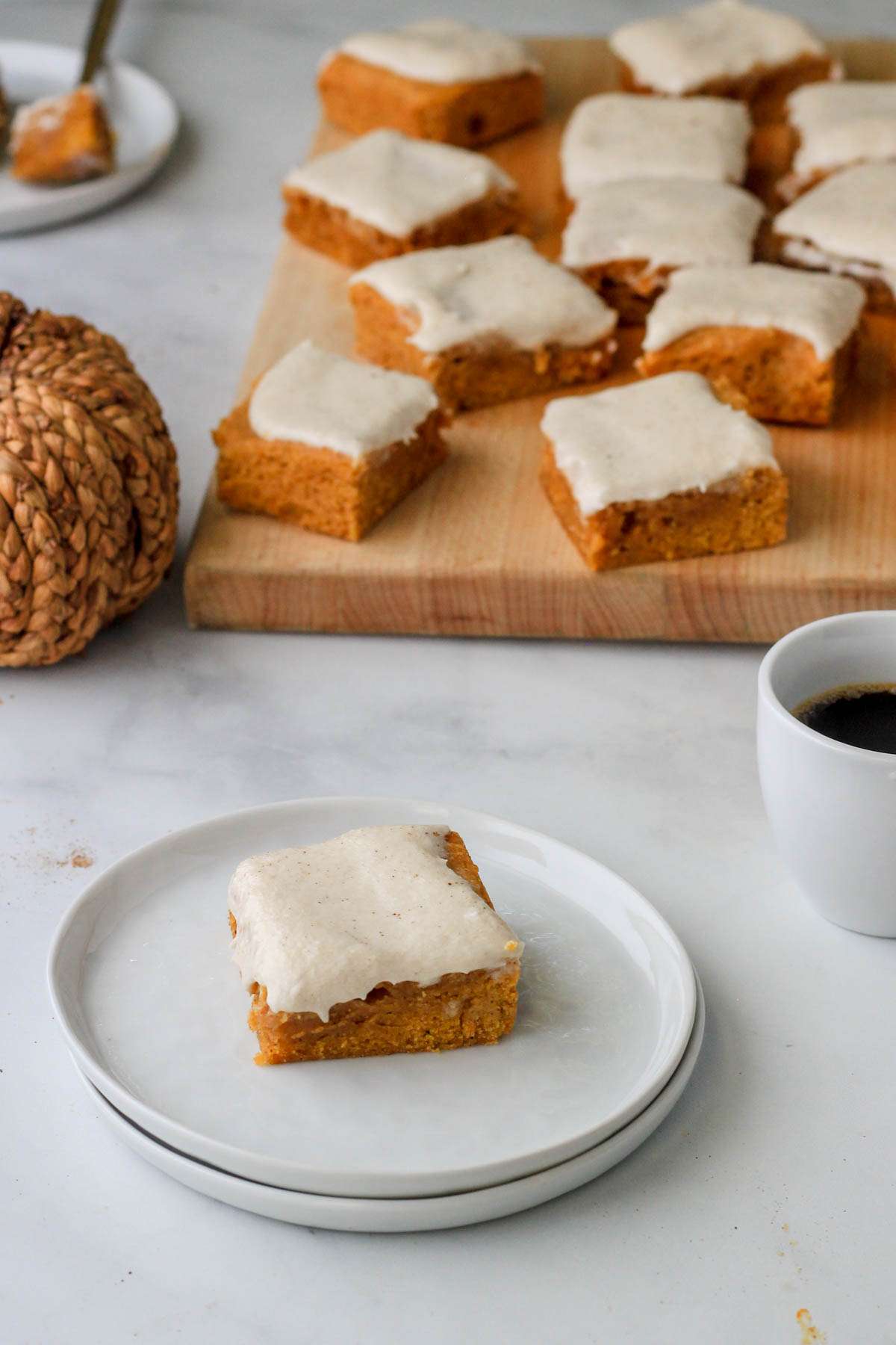 A small stack of plates topped with a dairy-free pumpkin spice cookie bar in front of a cup of coffee to the right, a cutting board of cookie bars in the back and a woven pumpkin to the left.