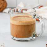 A vegan pumpkin spice latte with oat milk on a white counter in a glass mug.