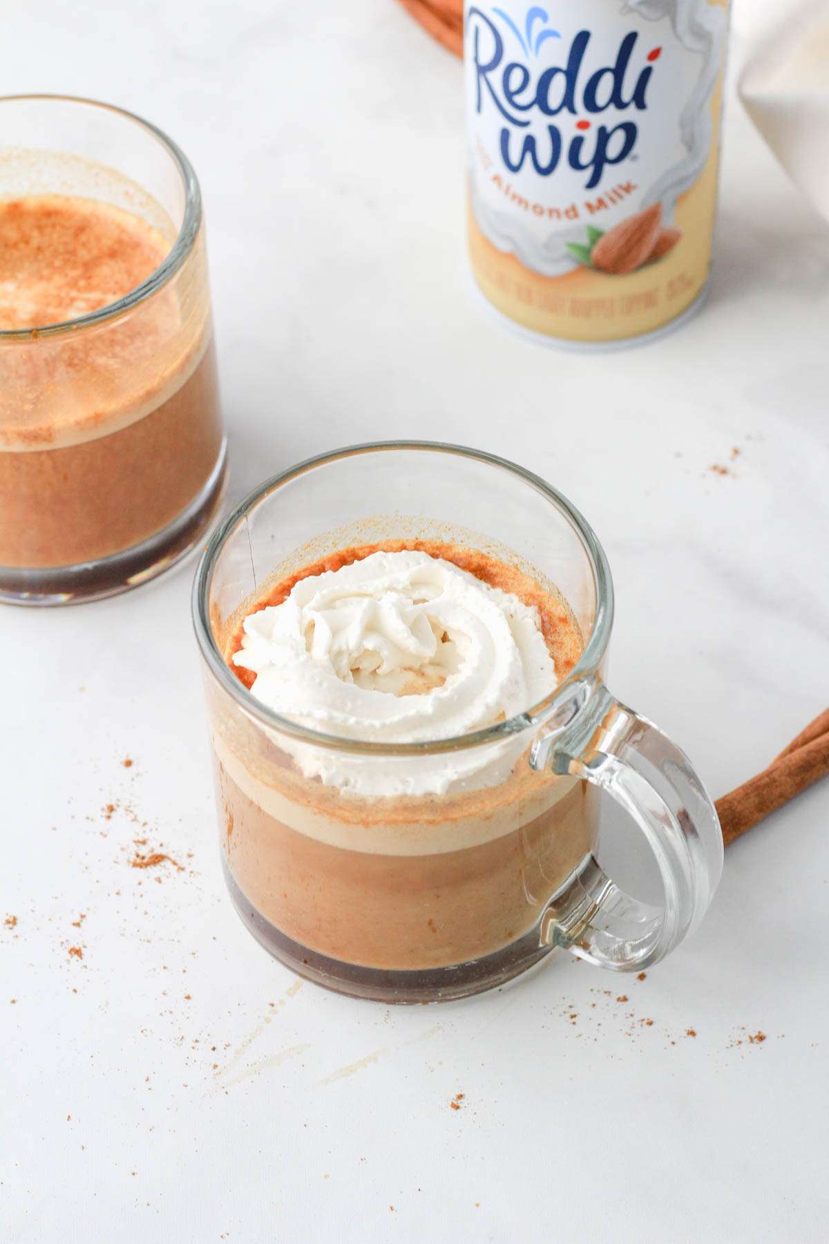 Non-dairy whipped topping on a vegan pumpkin spice latte with a cinnamon stick to the right of the cup.