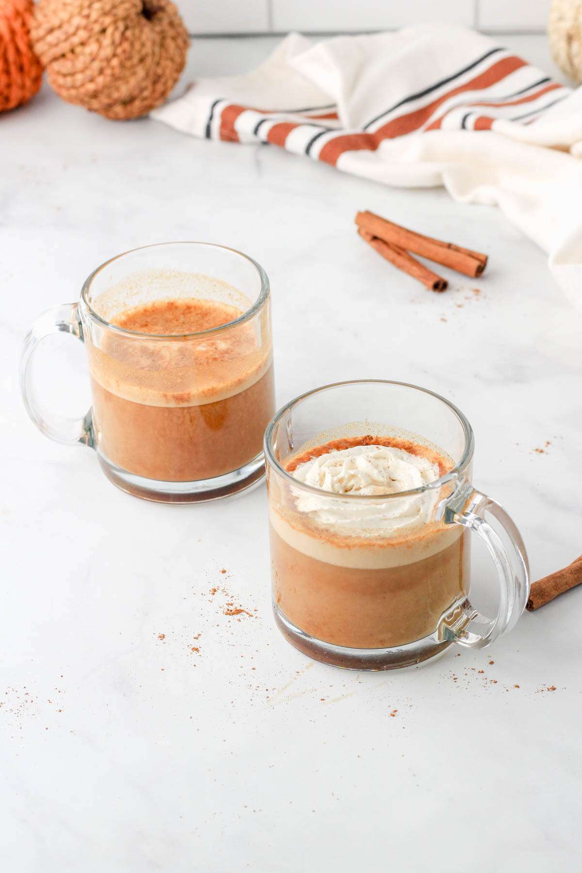 Two glass mugs with vegan pumpkin spice lattes made with oat milk on a white counter with two small pumpkins in the back left.