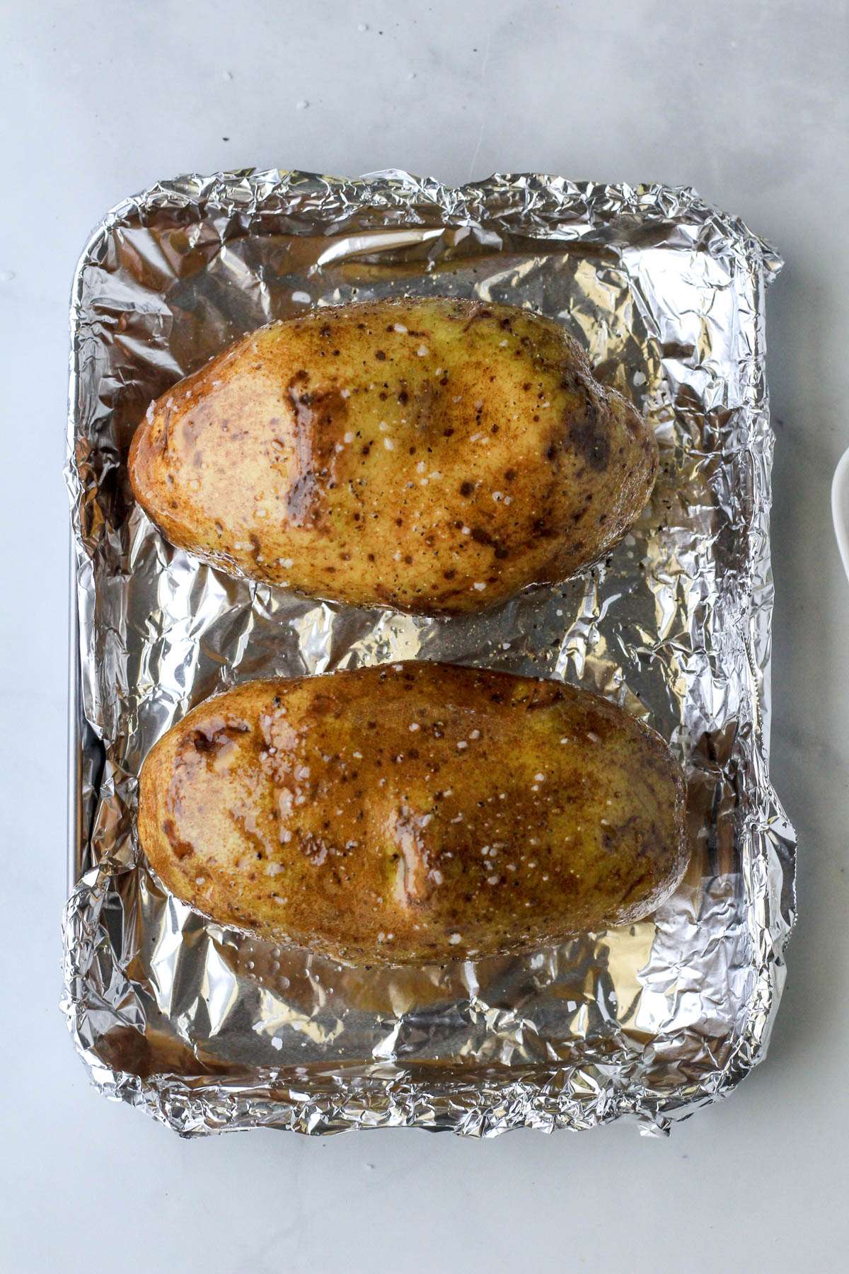 A small rimmed baking sheet lined with foil and topped with two baked potatoes dressed with oil, salt, and pepper before baking.