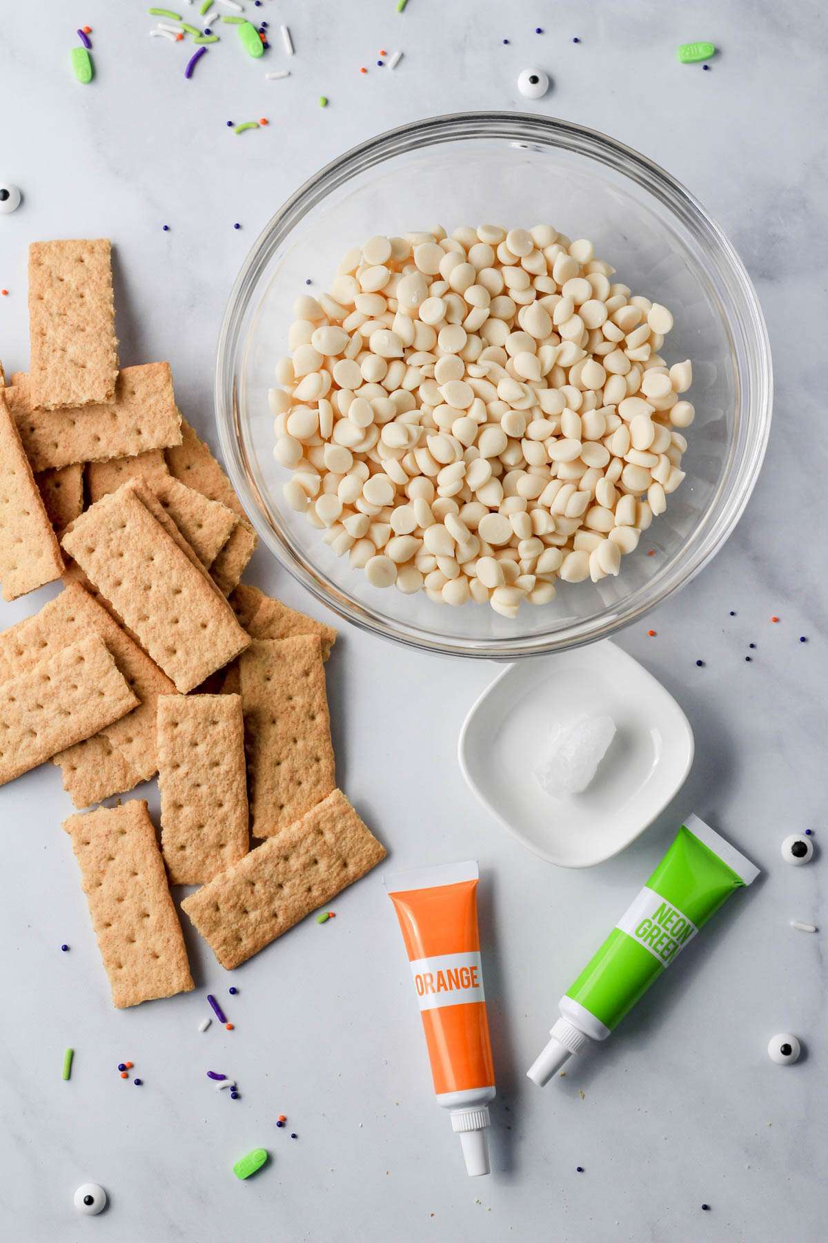 A glass bowl with dairy-free white chocolate next to graham crackers, coconut oil, and food coloring.