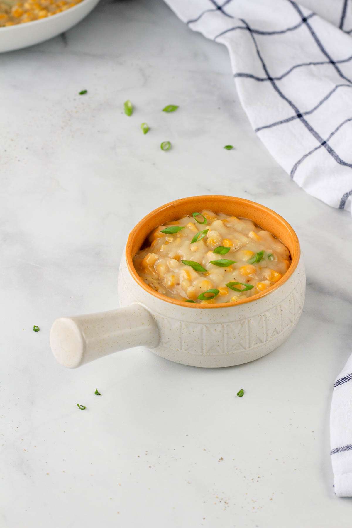 A small orange and white bowl filled with creamed corn topped with green onion on a white counter.