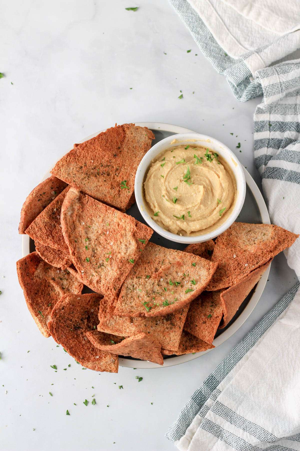 A green plate topped with air fryer pita chips and a small bowl of hummus in the top right side and a green and white dish towel on the right of the counter.