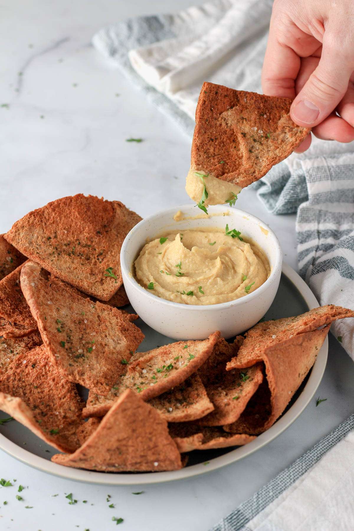 A hand holding a pita chip dipped in hummus over a bowl of hummus on a green plate of pita chips.