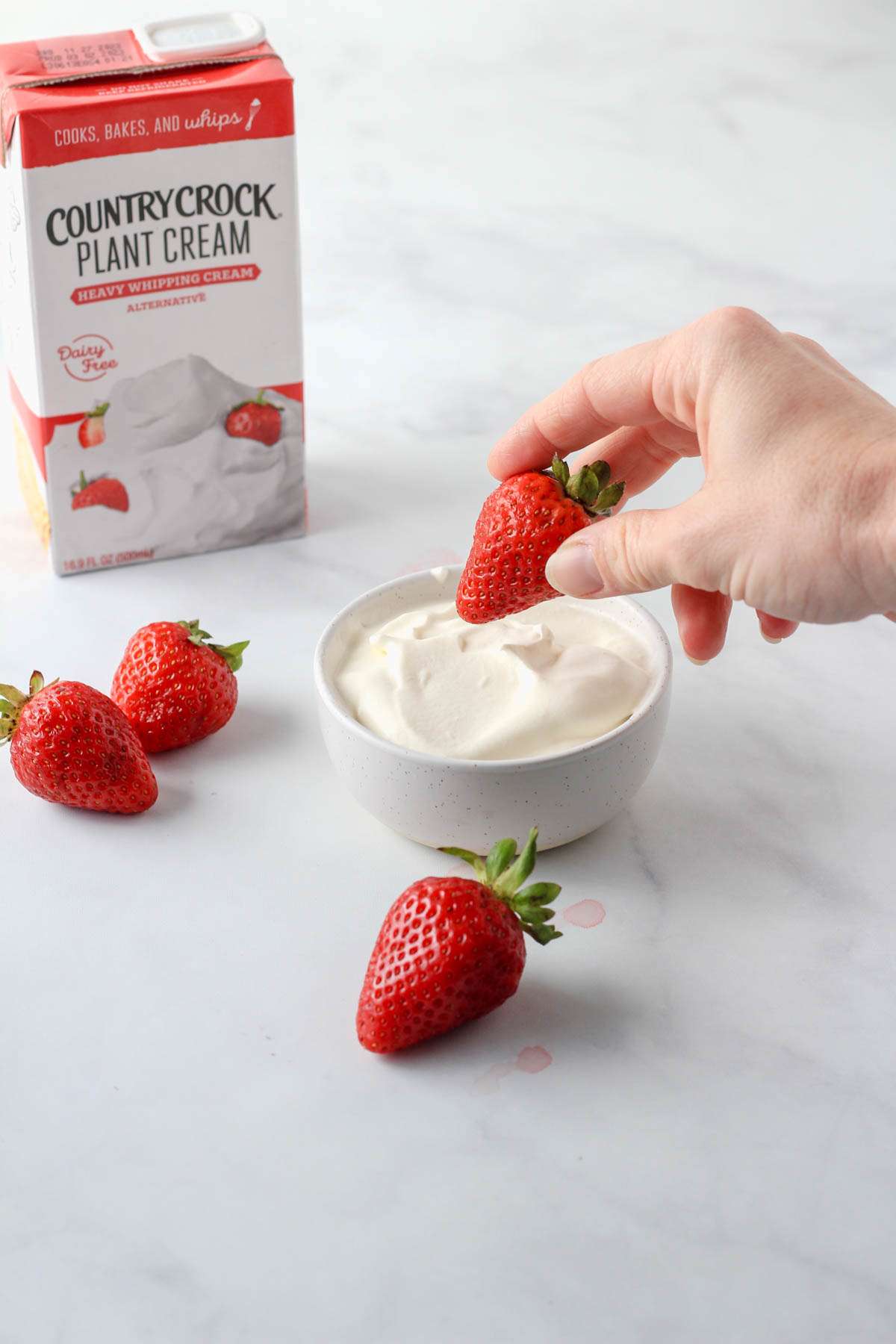 A hand holding a strawberry over a bowl of vegan whipped cream with a box of country crock plant cream in the back left.