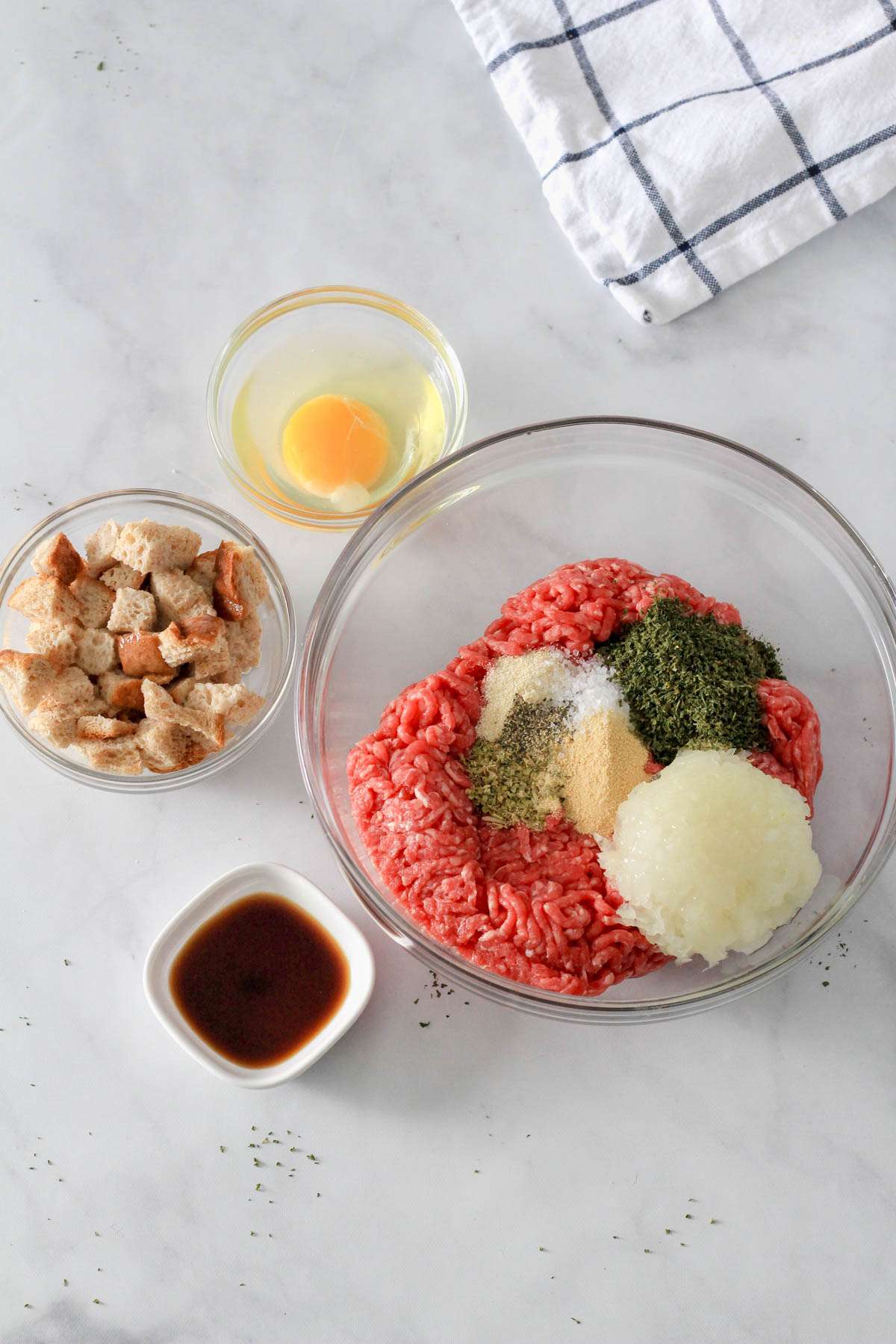 Glass bowls with ingredients for dairy-free meatball subs on a white counter before adding everything into the bowl with ground beef.