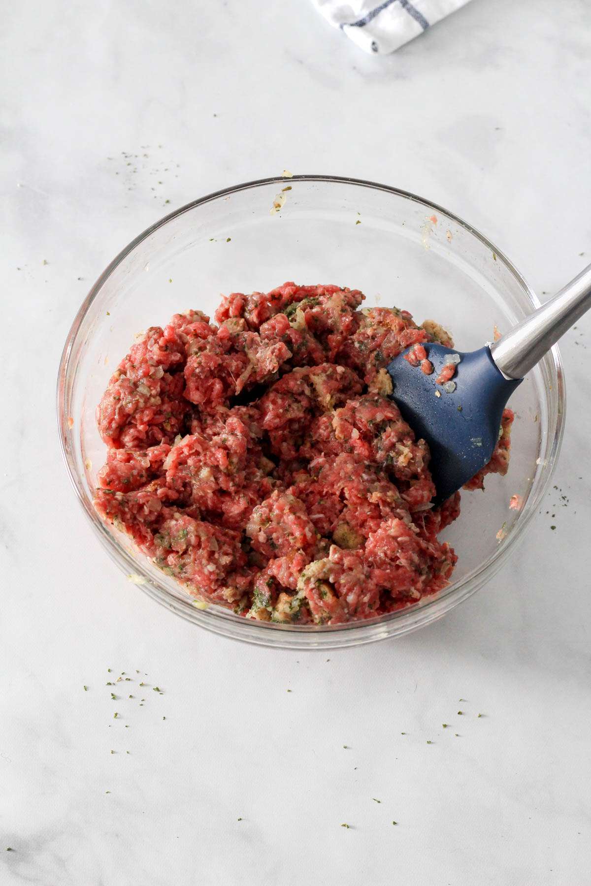 A glass bowl with meatball ingredients mixed together with a blue and silver spatula.