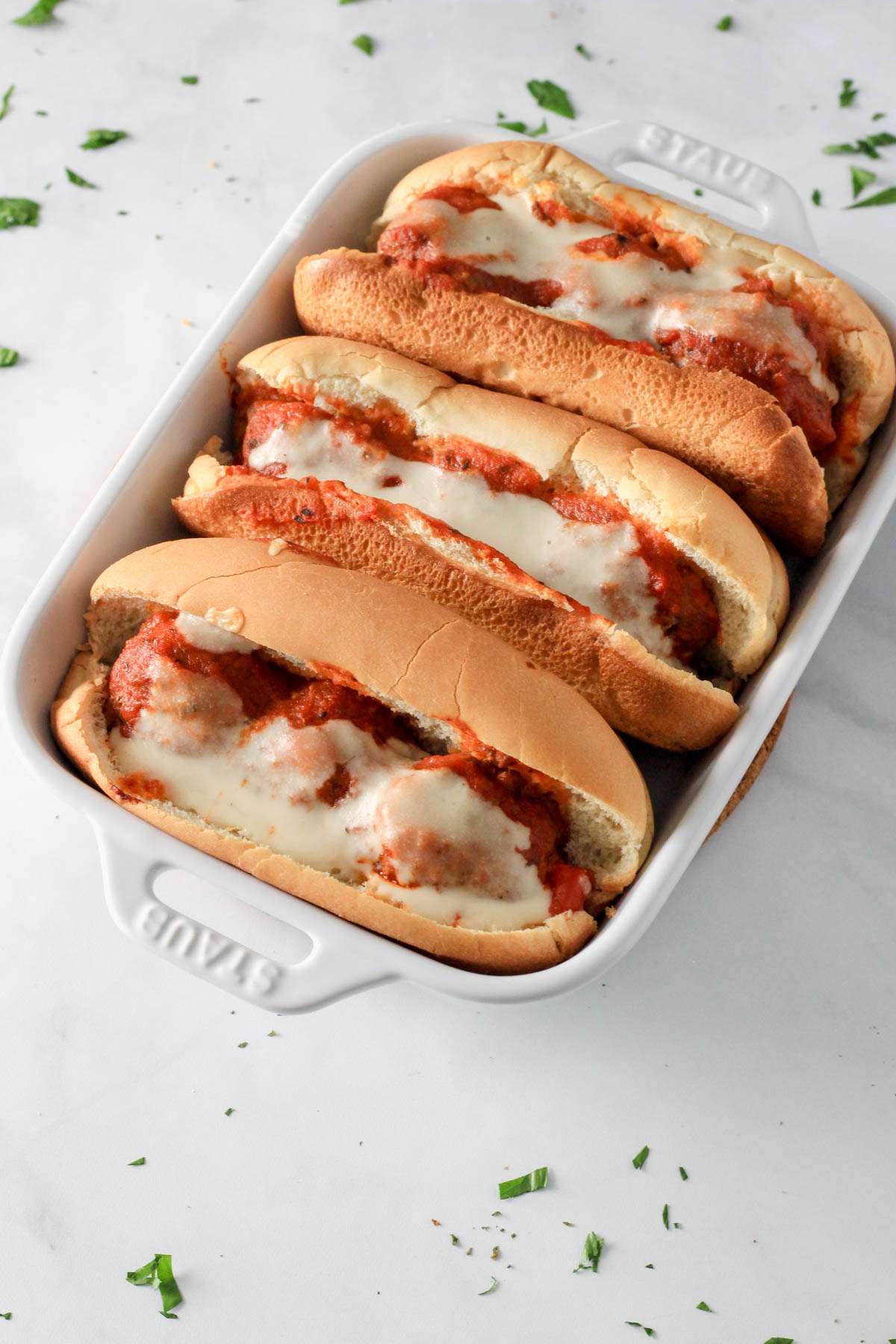Three baked dairy-free meatball subs in a white baking dish on a white counter with parsley.