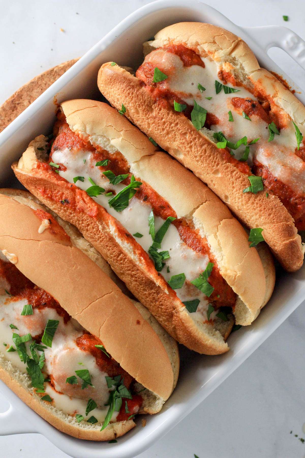Three dairy-free meatball subs in a white baking dish on a trivet with parsley sprinkled on top of the subs.