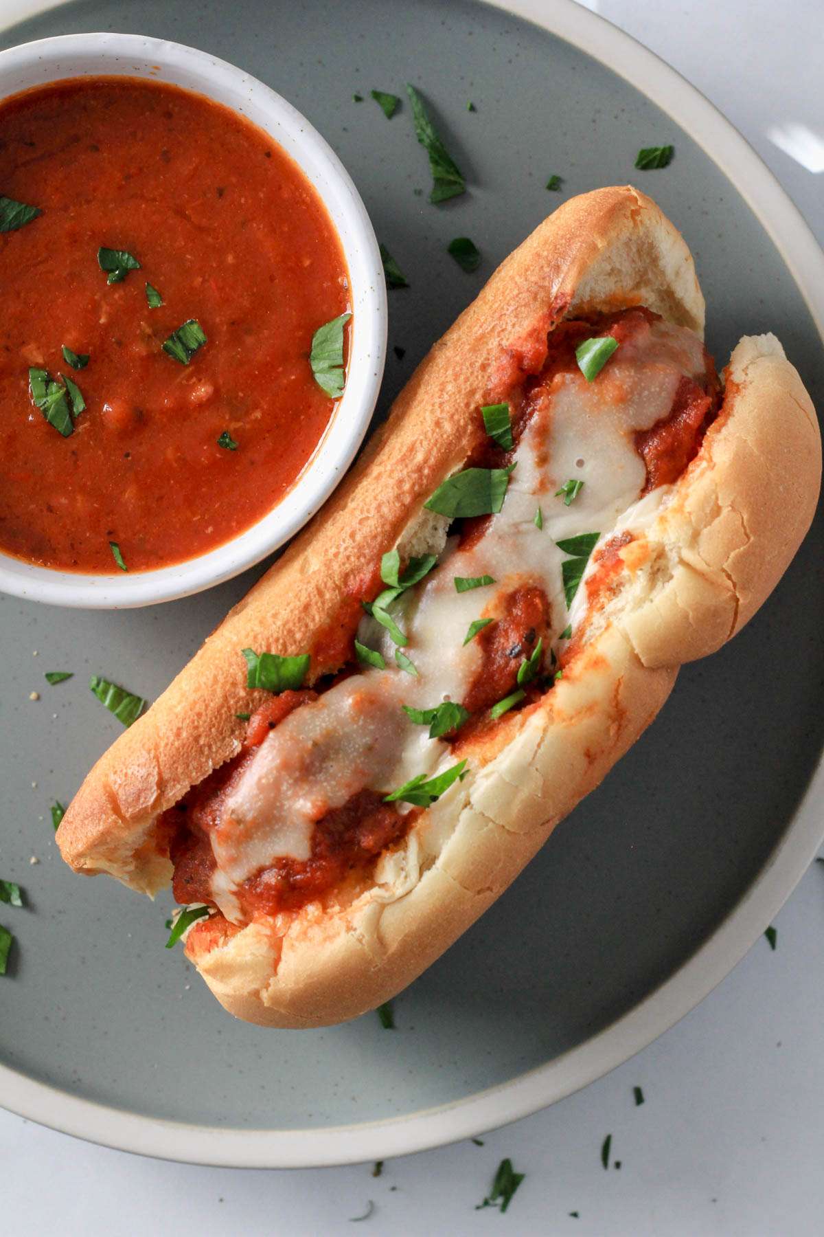 A green plate with a dairy-free meatball sub topped with parsley with a bowl of marinara sauce to the left.