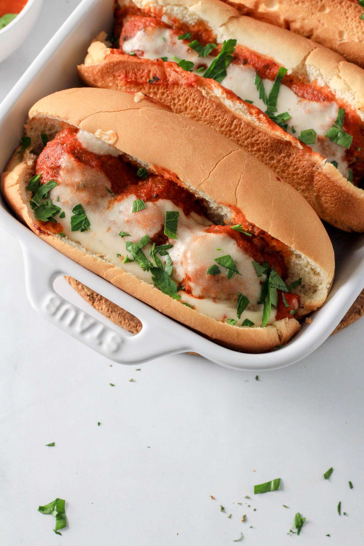 A white baking dish with a diary-free meatball sub topped with vegan cheese and parsley.