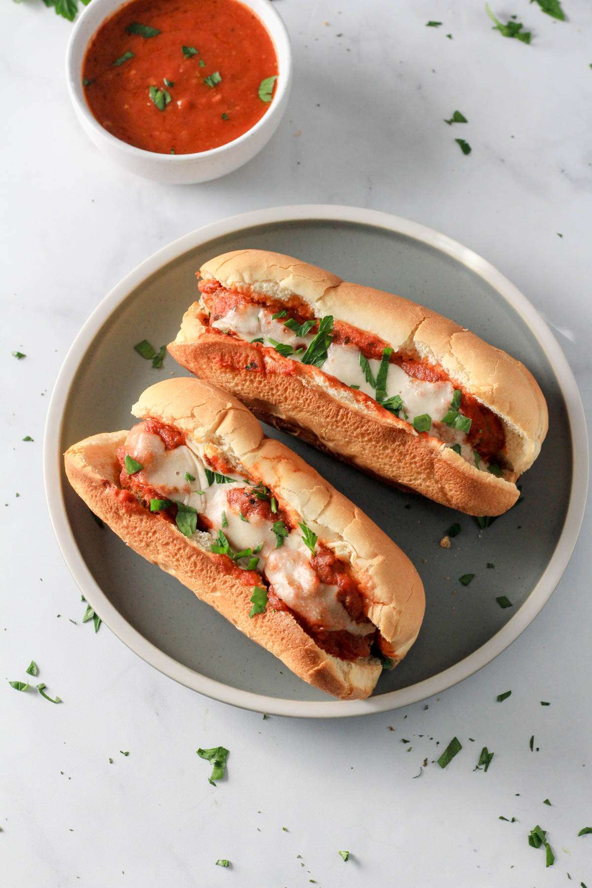 A green plate with two meatball subs topped with vegan cheese and parsley with a small white bowl of marinara in the top left.