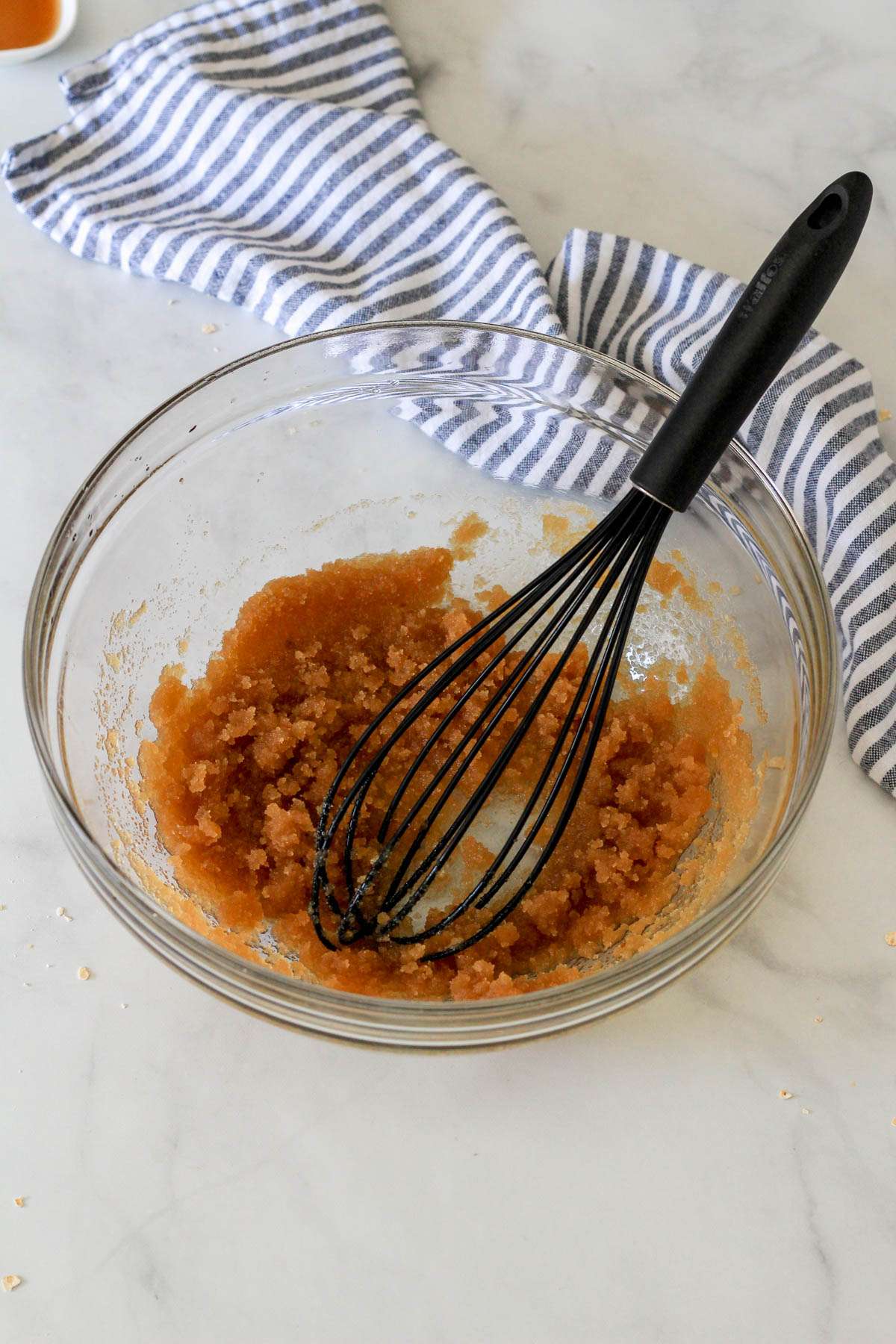 A glass bowl with brown sugar whisked with coconut oil and a black whisk in the bowl.