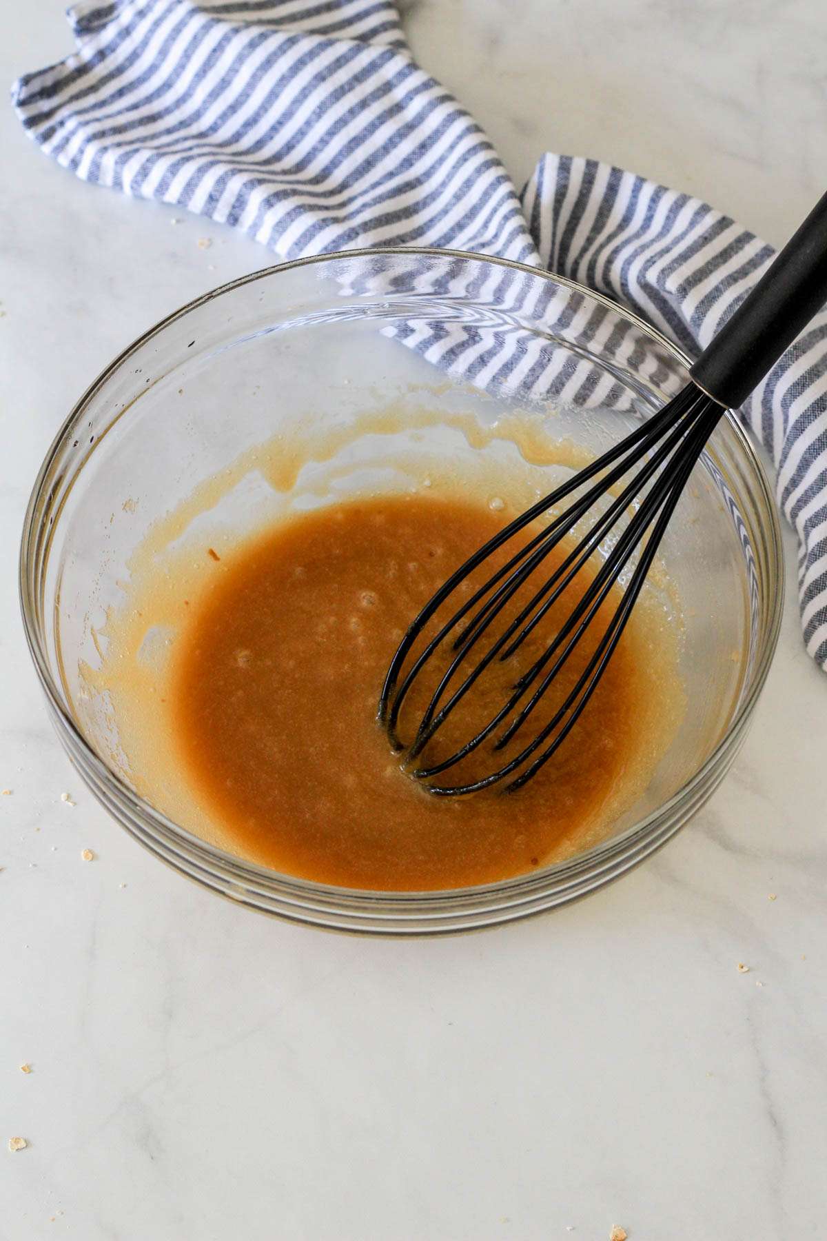 A glass mixing bowl with wet ingredients for dairy-free oatmeal chocolate chip cookie bars and a black whisk.