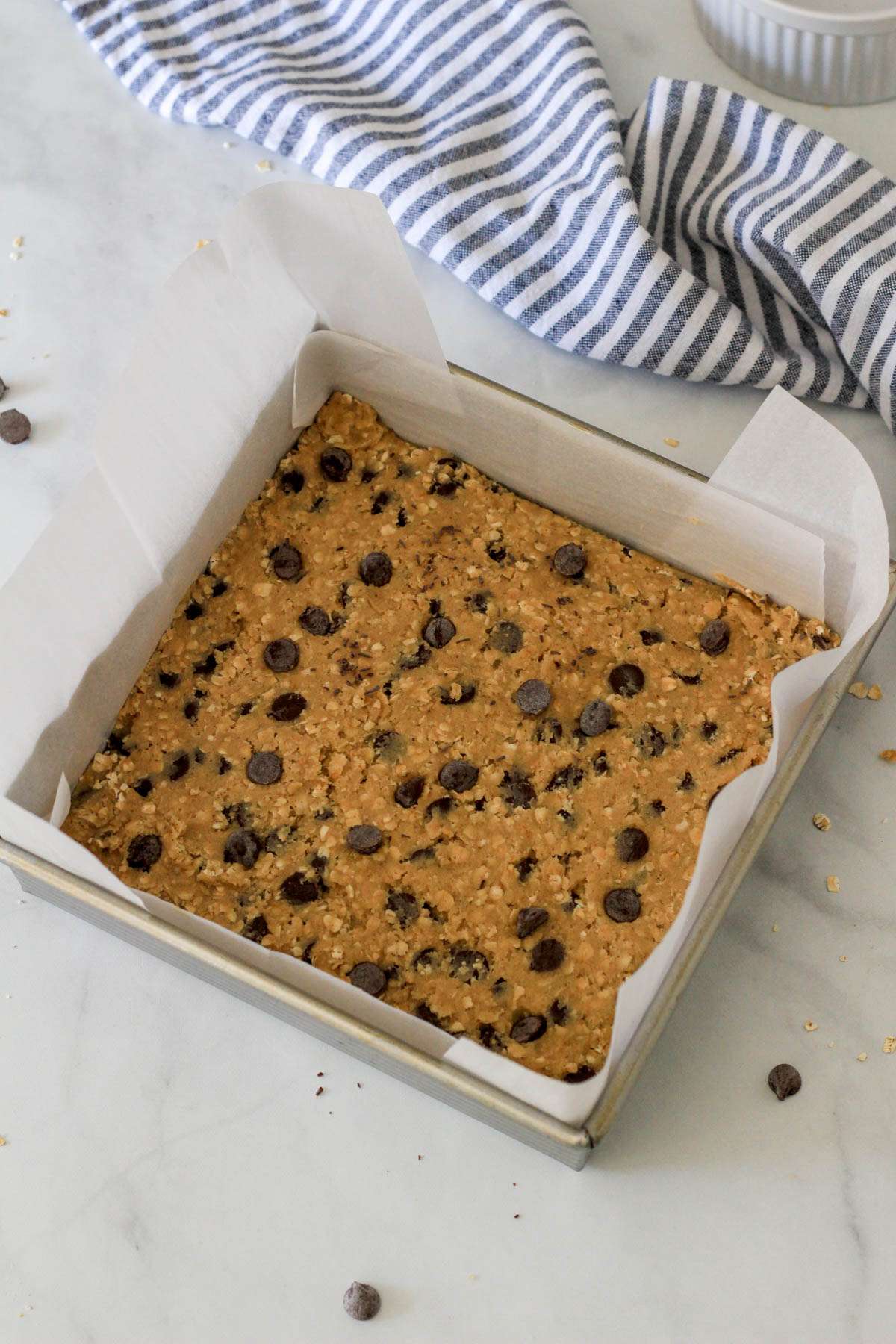 A metal baking pan lined with parchment paper with dairy-free oatmeal chocolate chip cookie bar dough before baking on a white counter with a blue and white striped towel in the top right.