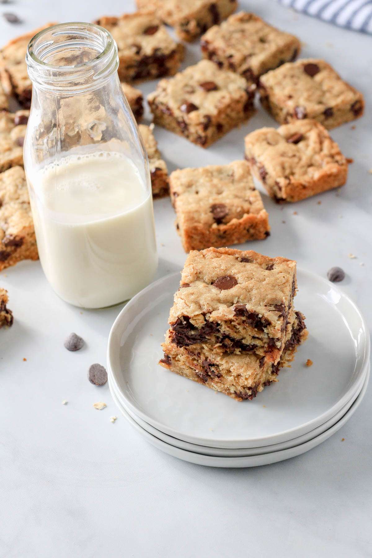 A small stack of white plates with two oatmeal chocolate chip cookie bars and a glass of non-dairy milk to the left with cookie bars in the back.