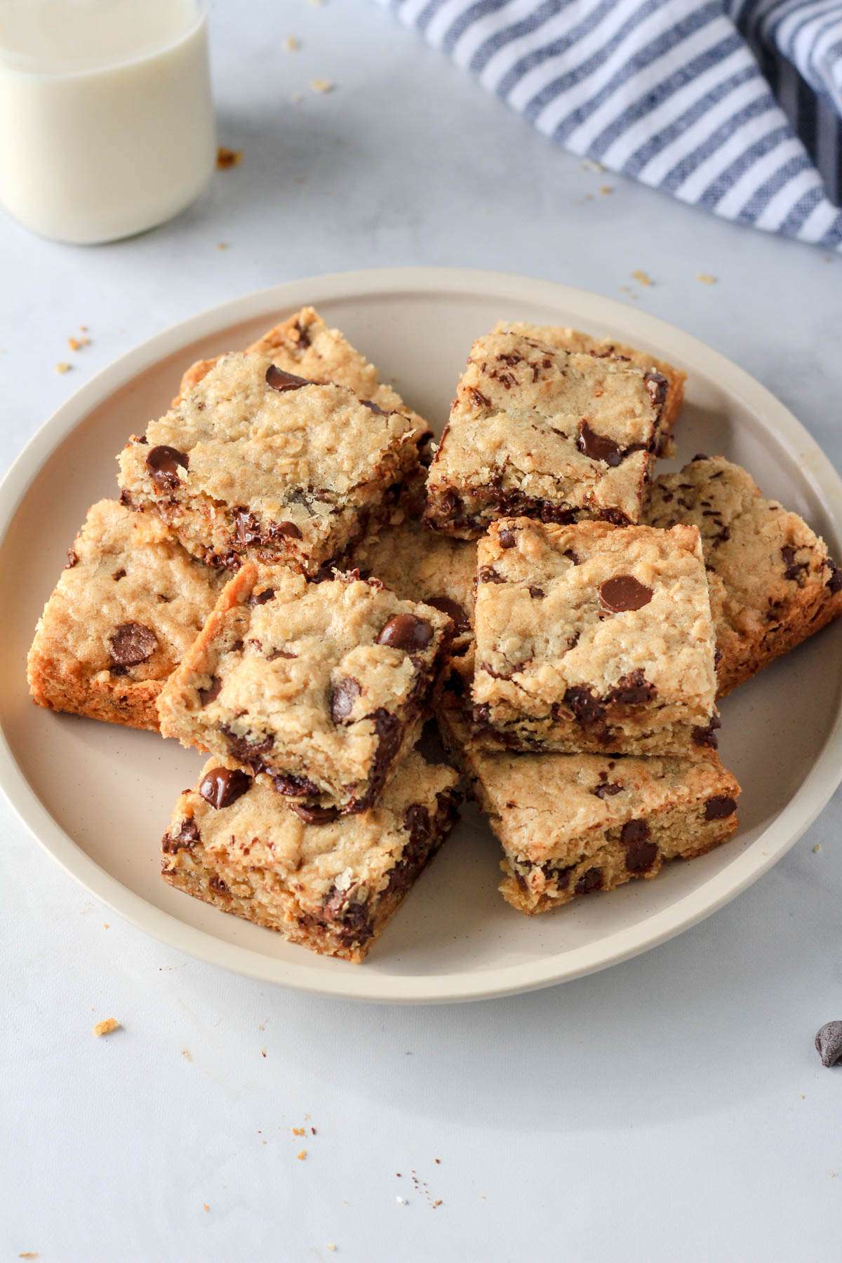 A cream plate of oatmeal chocolate chip cookie bars on a white counter with a glass of non-dairy milk in the back left.