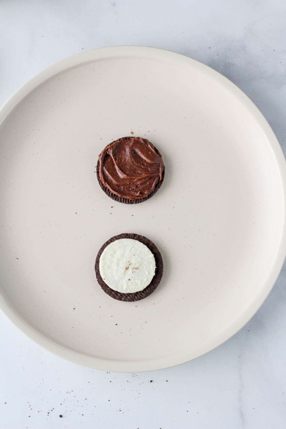 An opened Oreo on a white plate with the top part iced with chocolate frosting and the bottom part iced with Oreo creme.