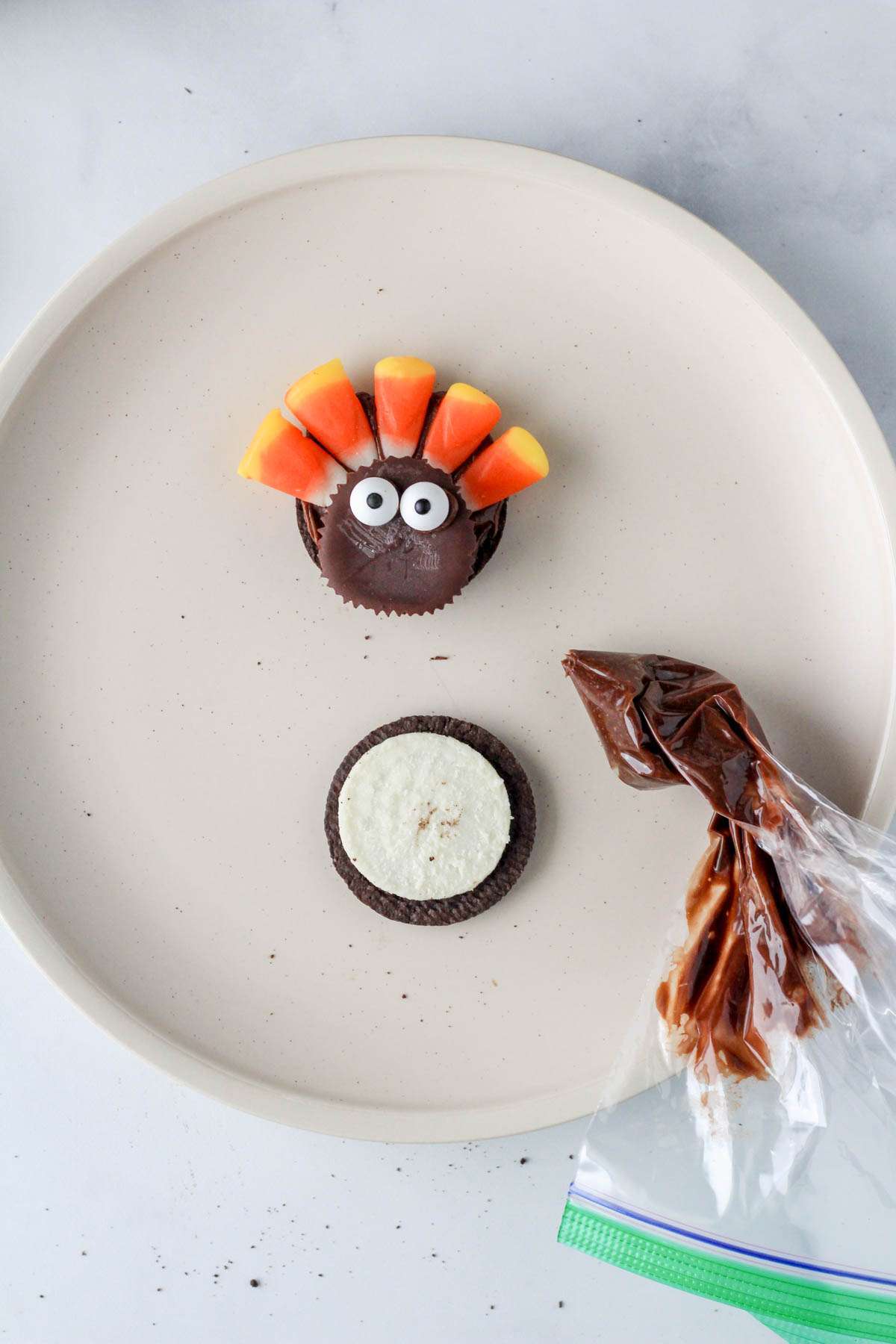 A piping bag of chocolate frosting on a white plate with a turkey made out of oreo, sun butter cups, candy corn, and candy eyes on top of a white plate.