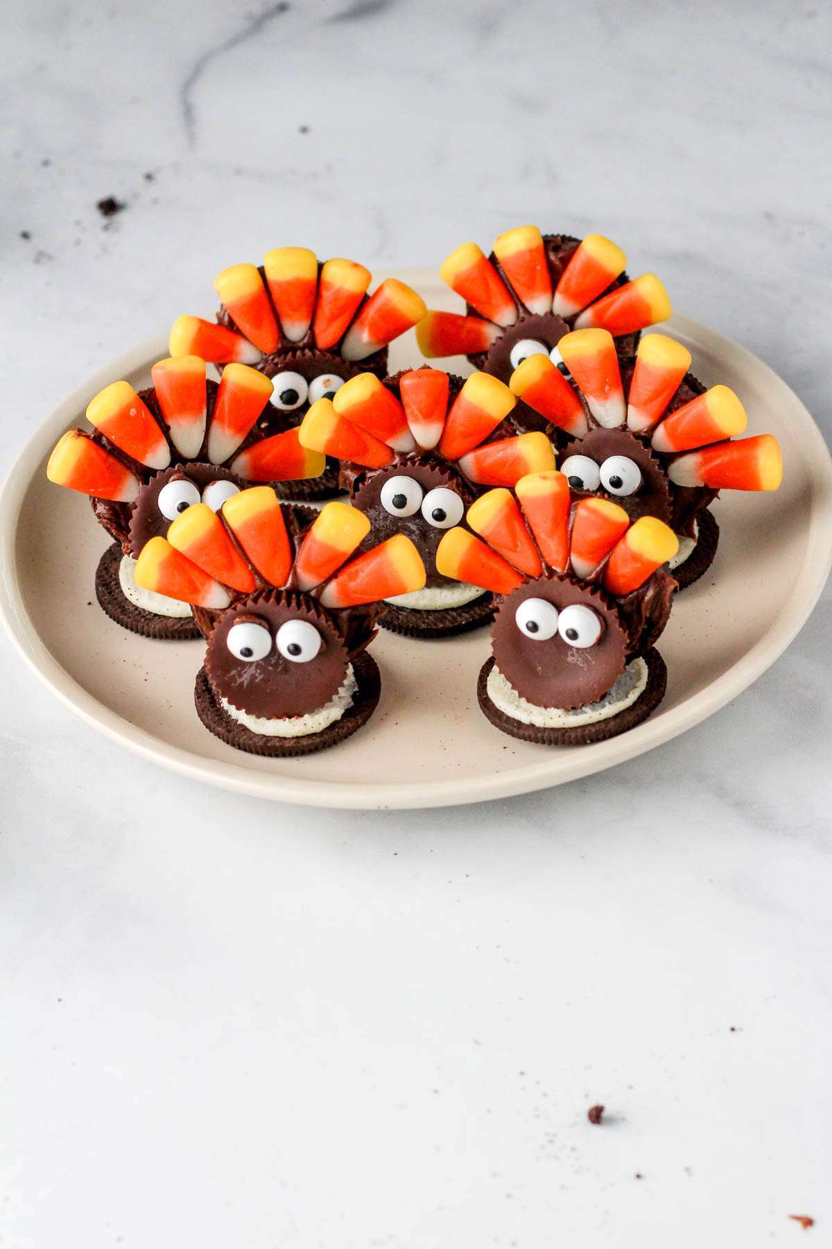 A white plate on a white counter with seven Oreo cookies made to look like turkeys using sun butter cups, candy corn, and candy eyes.