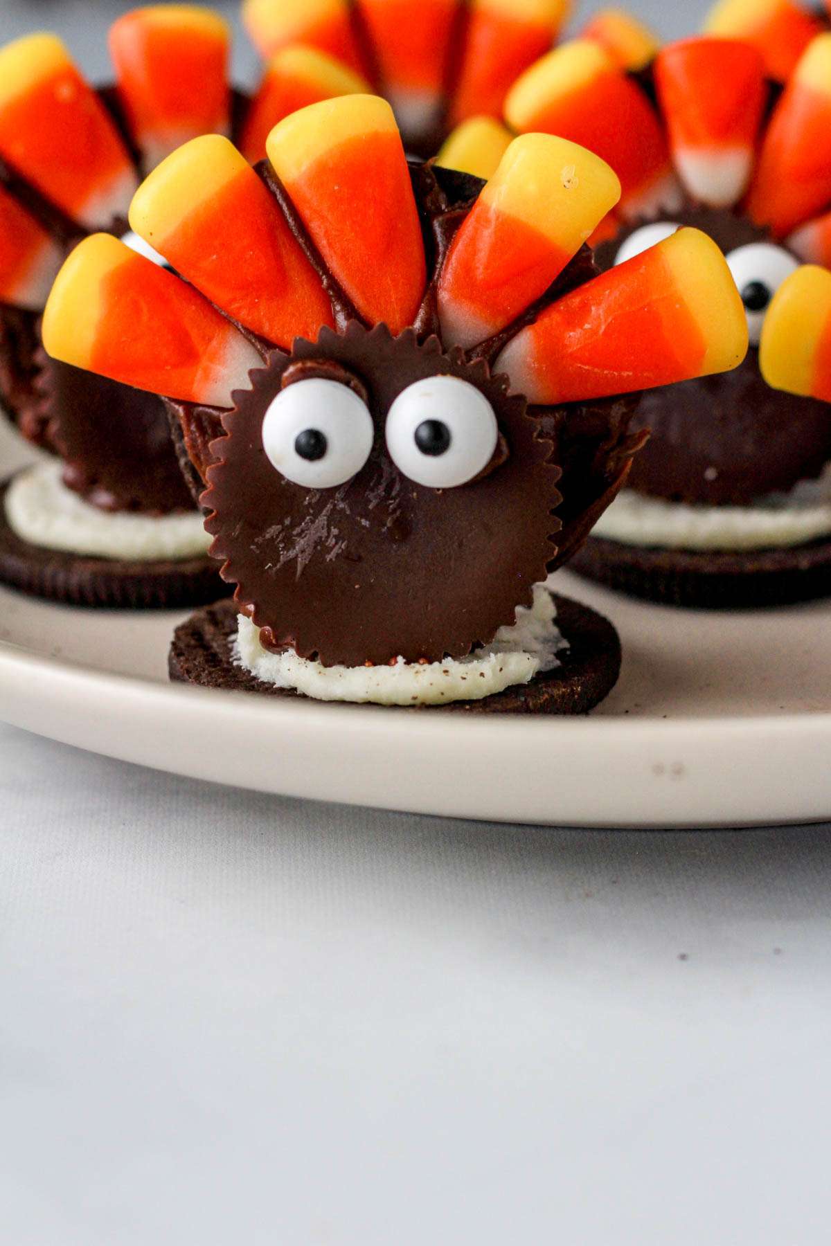 A close up of a turkey Oreo cookie on a white plate on a white counter with additional turkeys behind it.