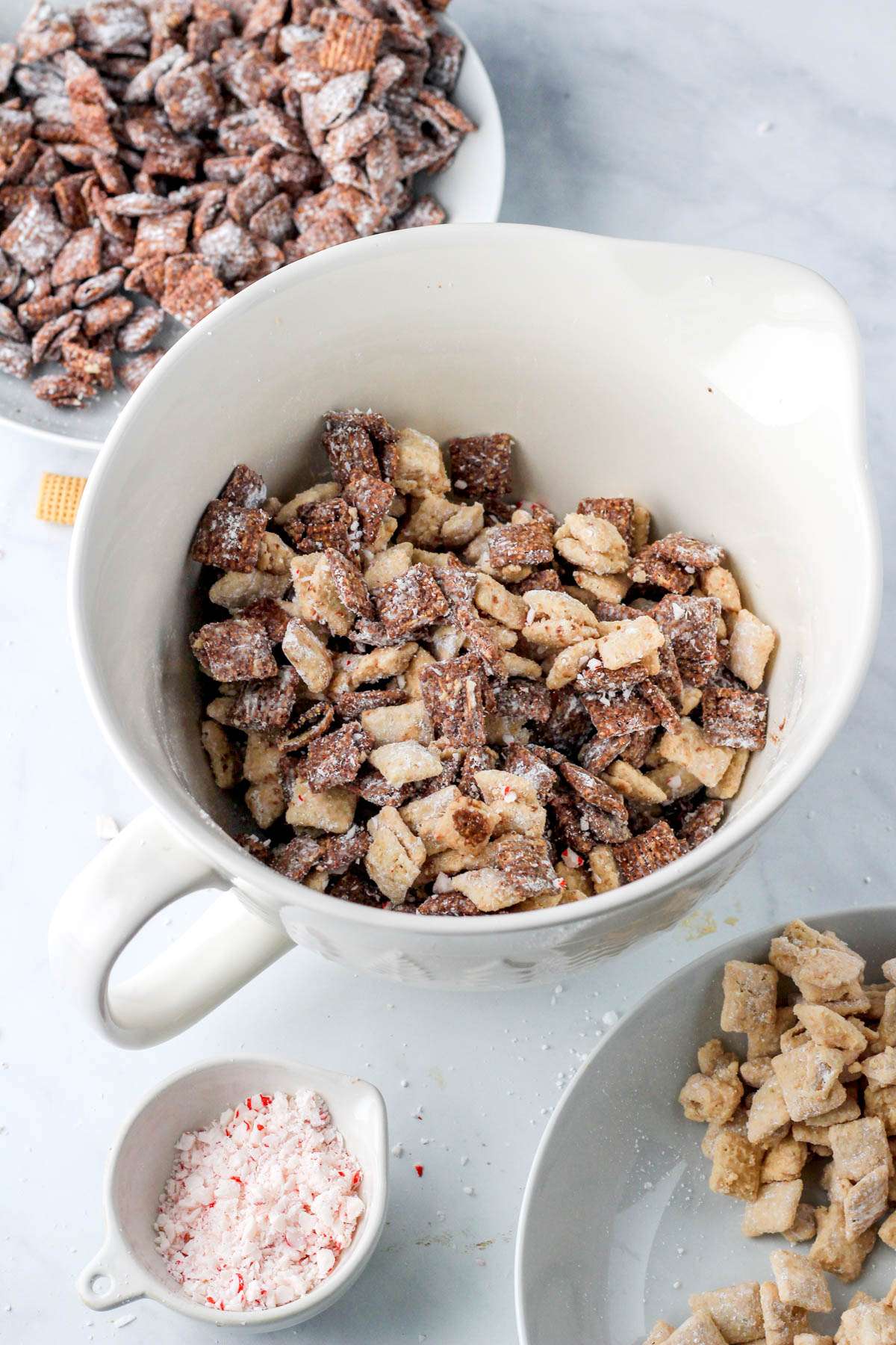 A white mixing bowl with a mixture of peppermint bark puppy chow and a dark chocolate puppy chow in the top left corner and a white chocolate puppy chow in the bottom right corner.