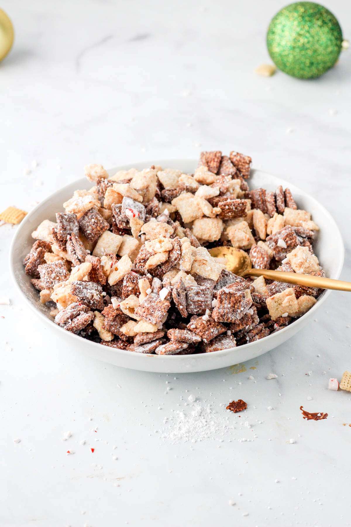 A white bowl of vegan peppermint bark puppy chow on a white counter with a green ornament in the back right.