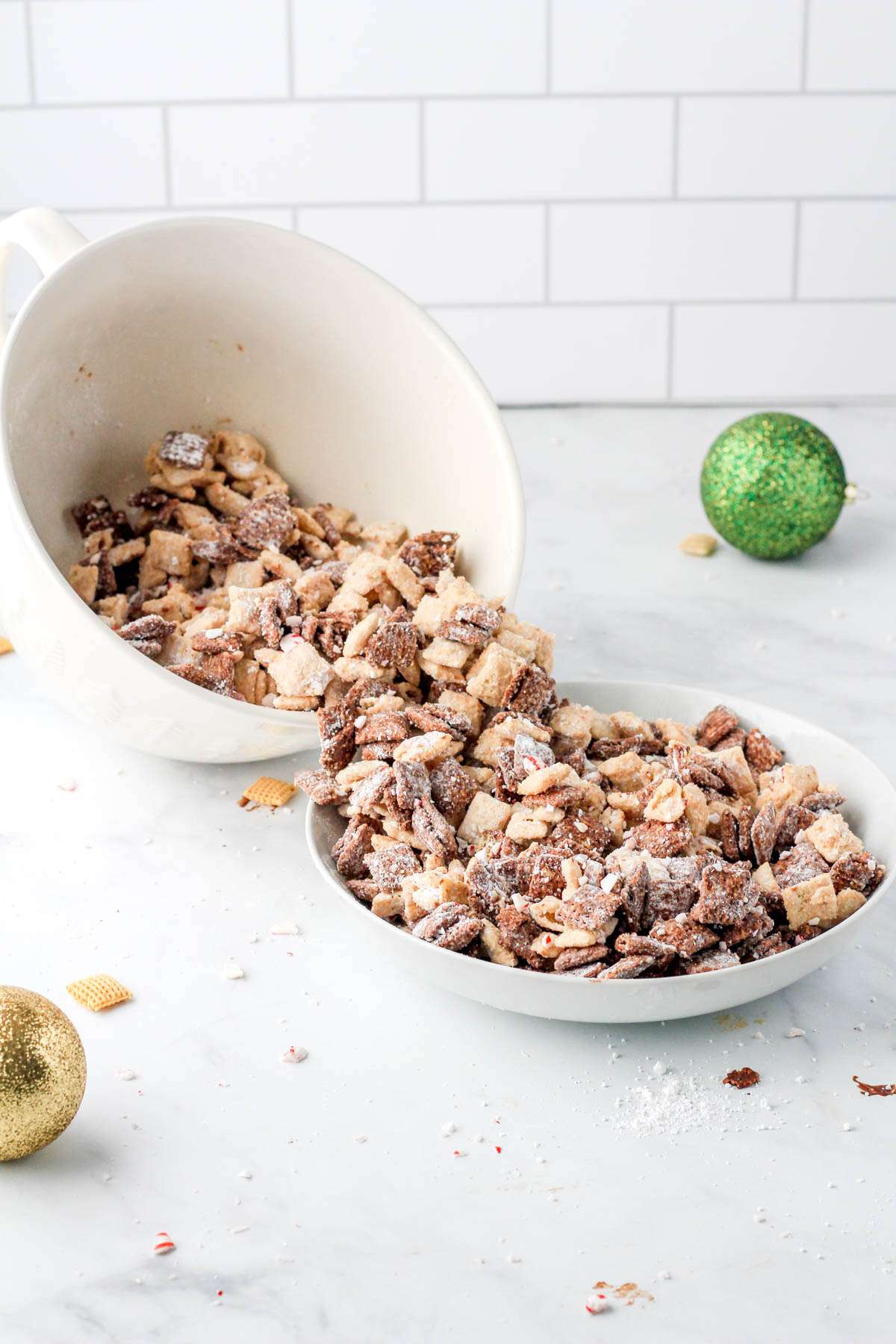 A large bowl of peppermint bark puppy chow pouring into a smaller bowl of puppy chow on a white counter with a green ornament in the back right corner.