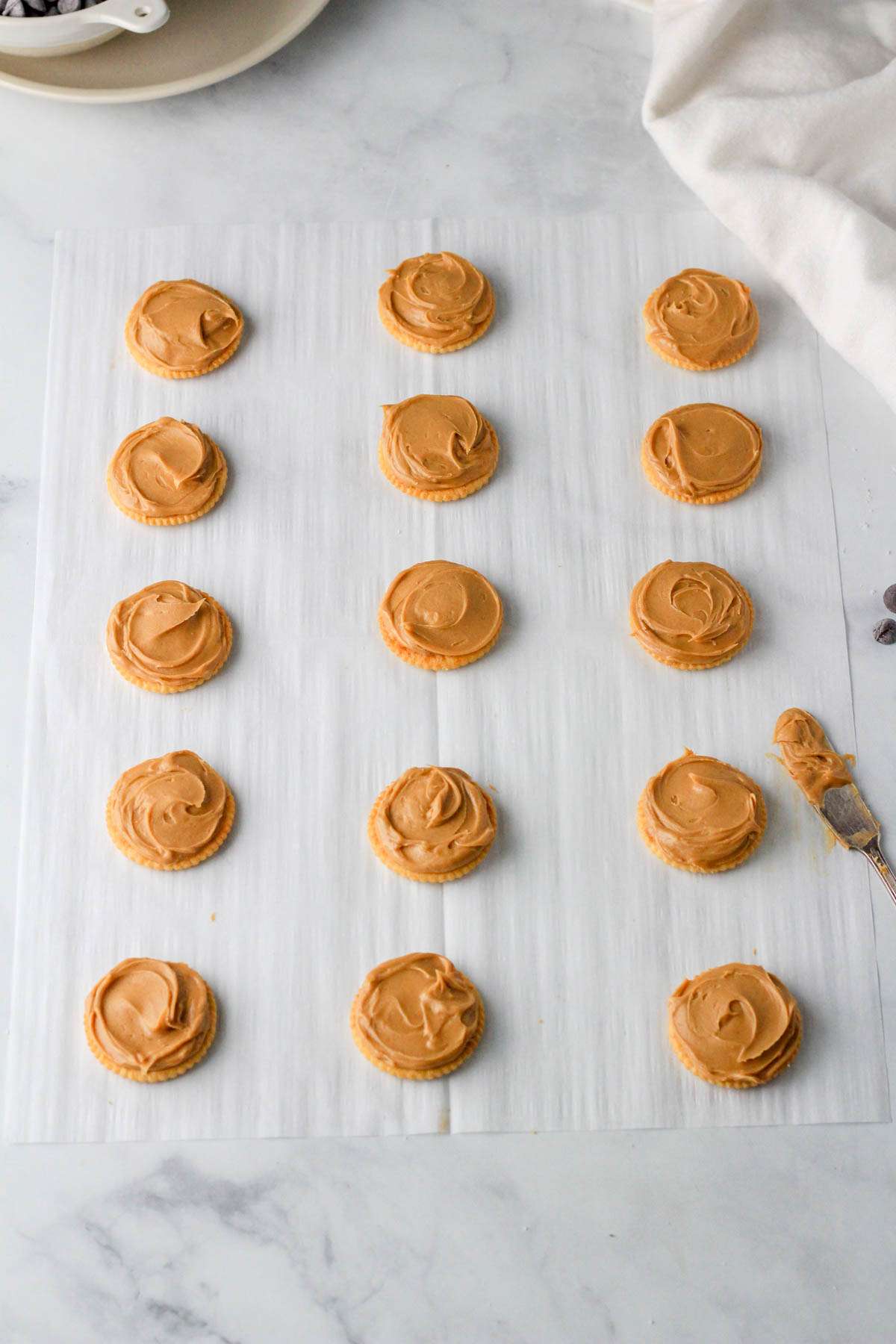 A parchment paper lined with Ritz crackers top side down and a half tablespoon of peanut butter spread on each cracker.