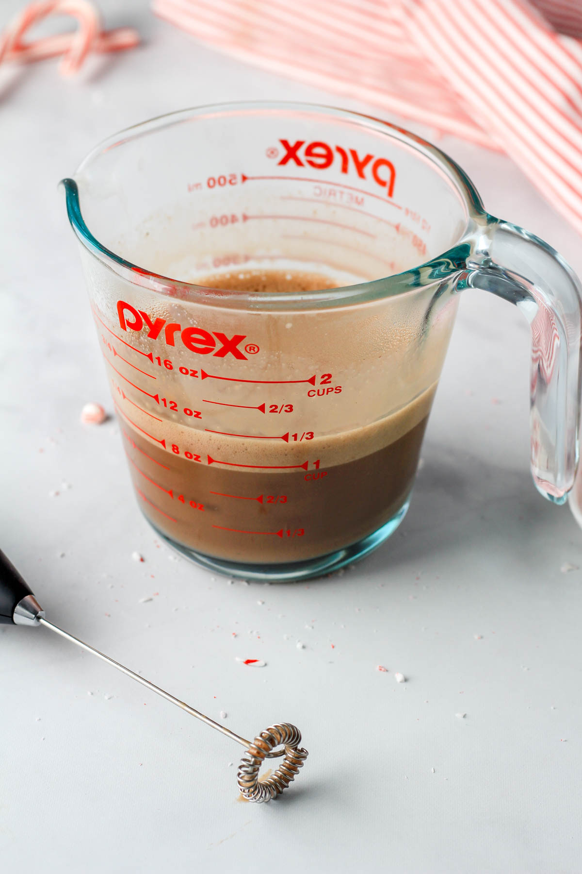A hand held frother in front of peppermint chocolate oat milk after frothing it in a liquid measuring cup.