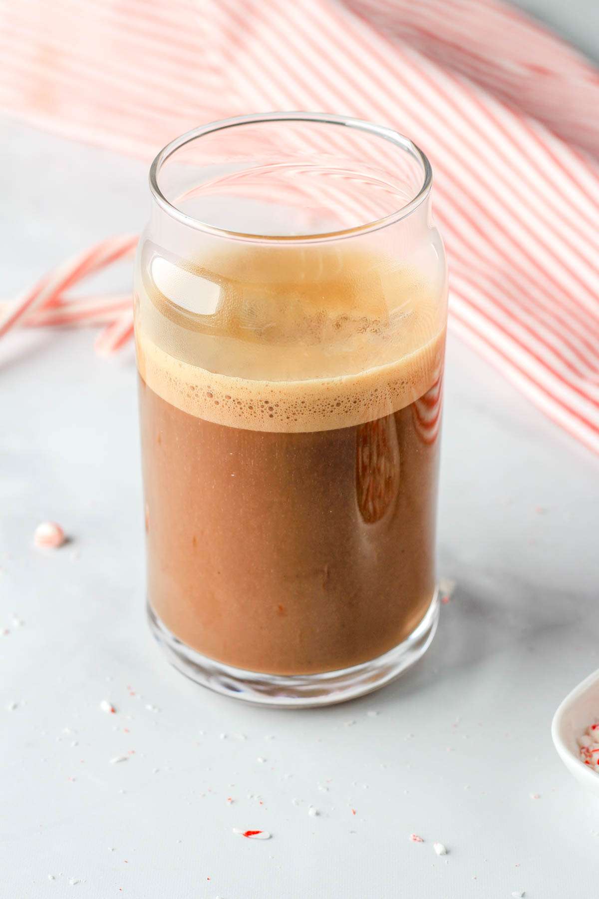 A glass cup with a vegan oat milk peppermint mocha on a white counter in front of a red and white striped dish towel.