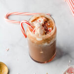 A glass cup with a vegan peppermint mocha topped with dairy-free whipped topping and crushed peppermint with a candy cane on the left side of the glass.