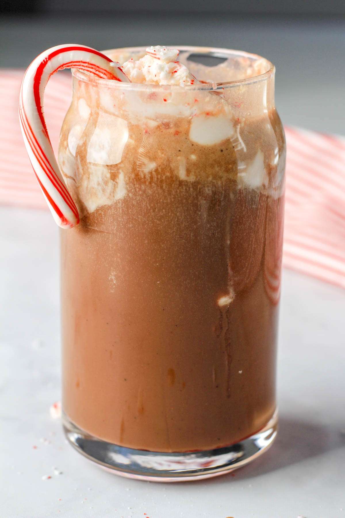 A close up of a glass with a vegan oat milk peppermint mocha topped with non-dairy whipped topping and a candy cane.