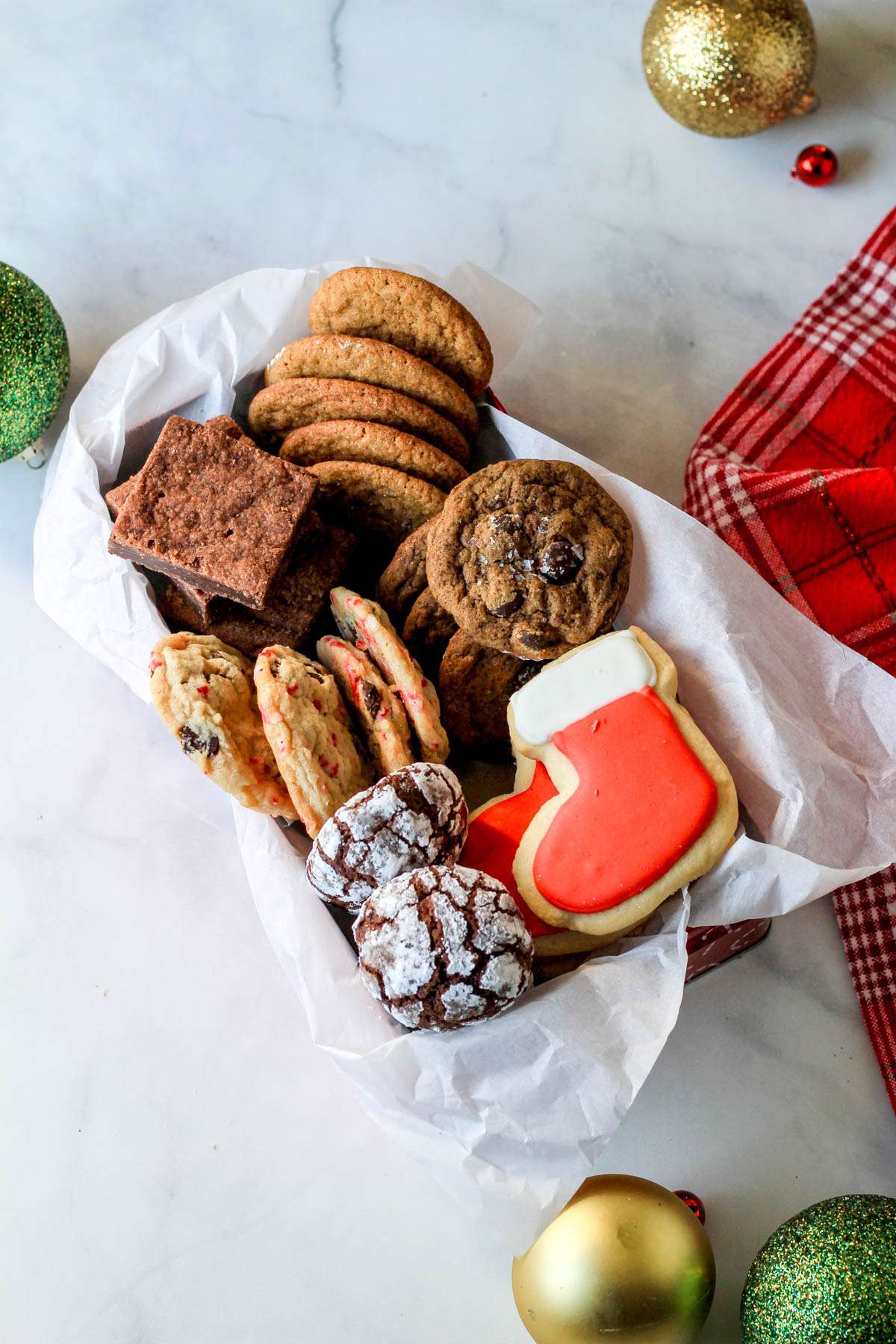 A parchment paper lined box with different holiday cookies on a white counter with green, gold, and red ornaments.