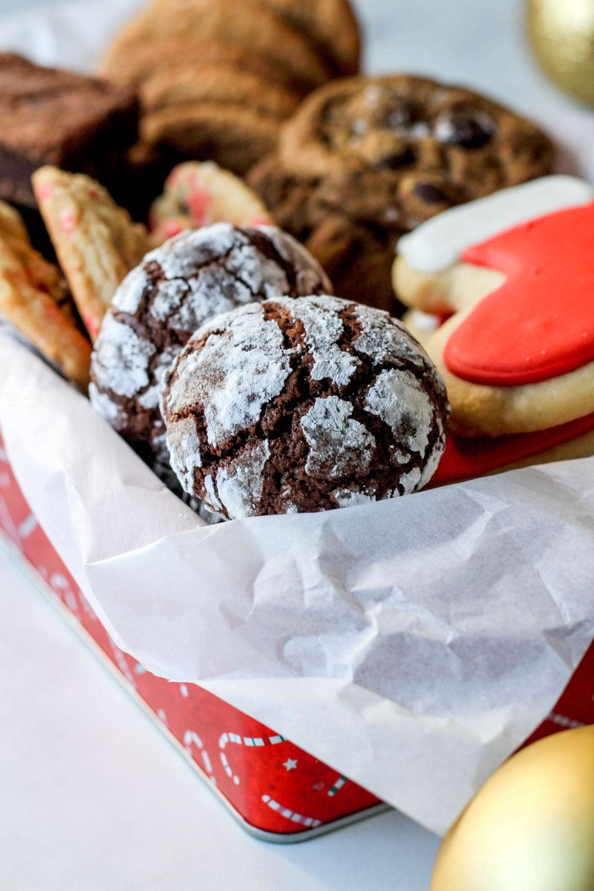 A red box lined with parchment paper from the side with chocolate crinkle cookies dusted in powdered sugar.