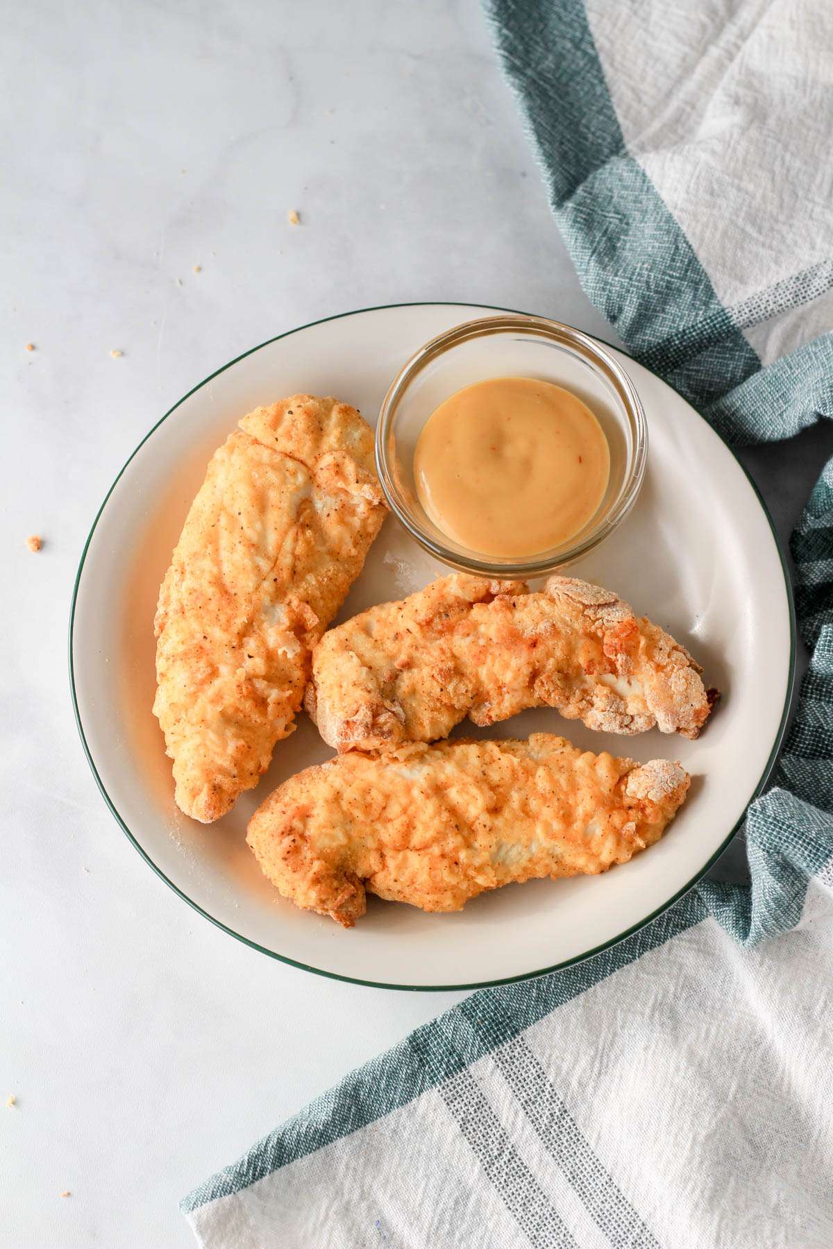 A white plate with a green rim and a small bowl of chick-fil-a sauce next to three air fryer chicken tenders.