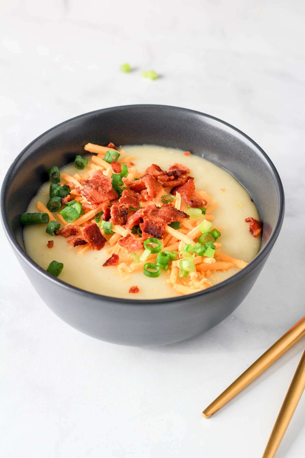 A navy bowl filled with dairy-free potato soup topped with bacon, shredded vegan cheese, and green onion with a gold spoon to the right.