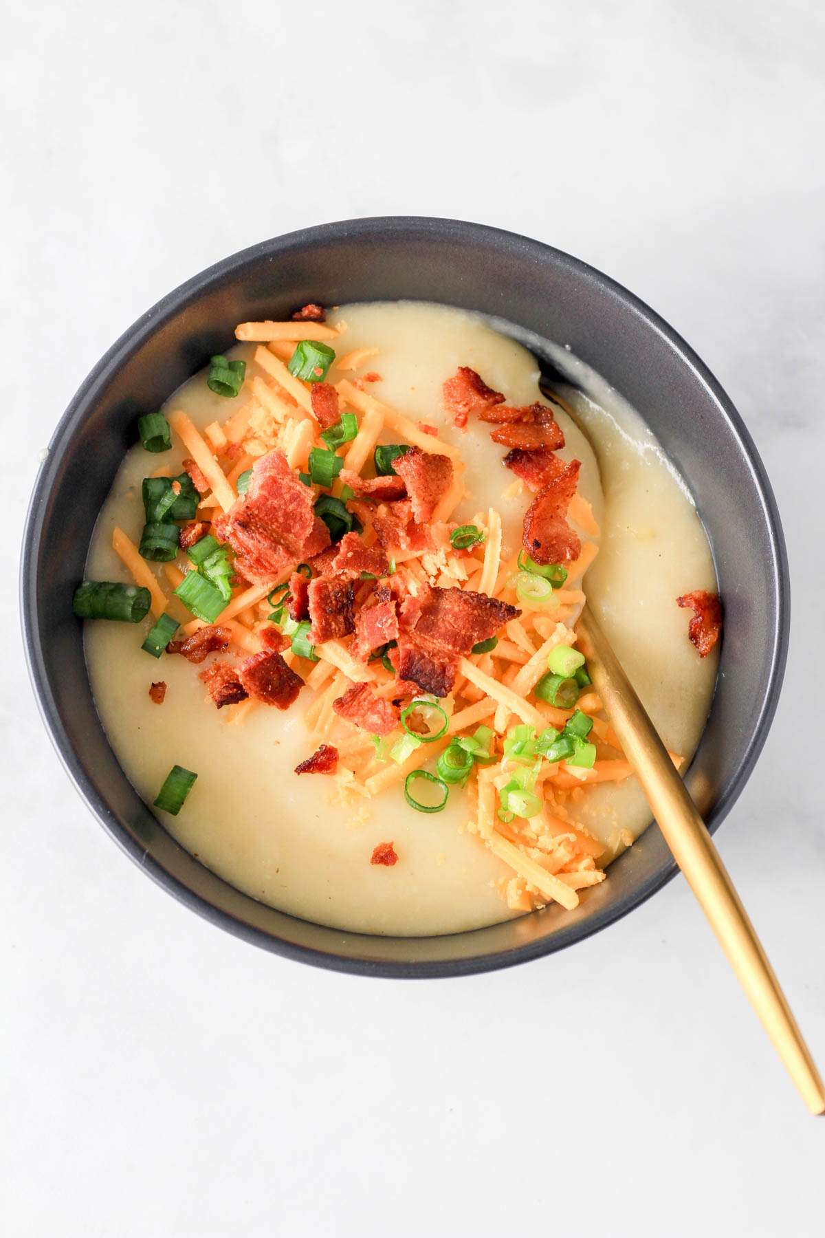 A top down image of dairy-free potato soup in a blue bowl topped with bacon, green onion, and vegan cheese on a white counter.