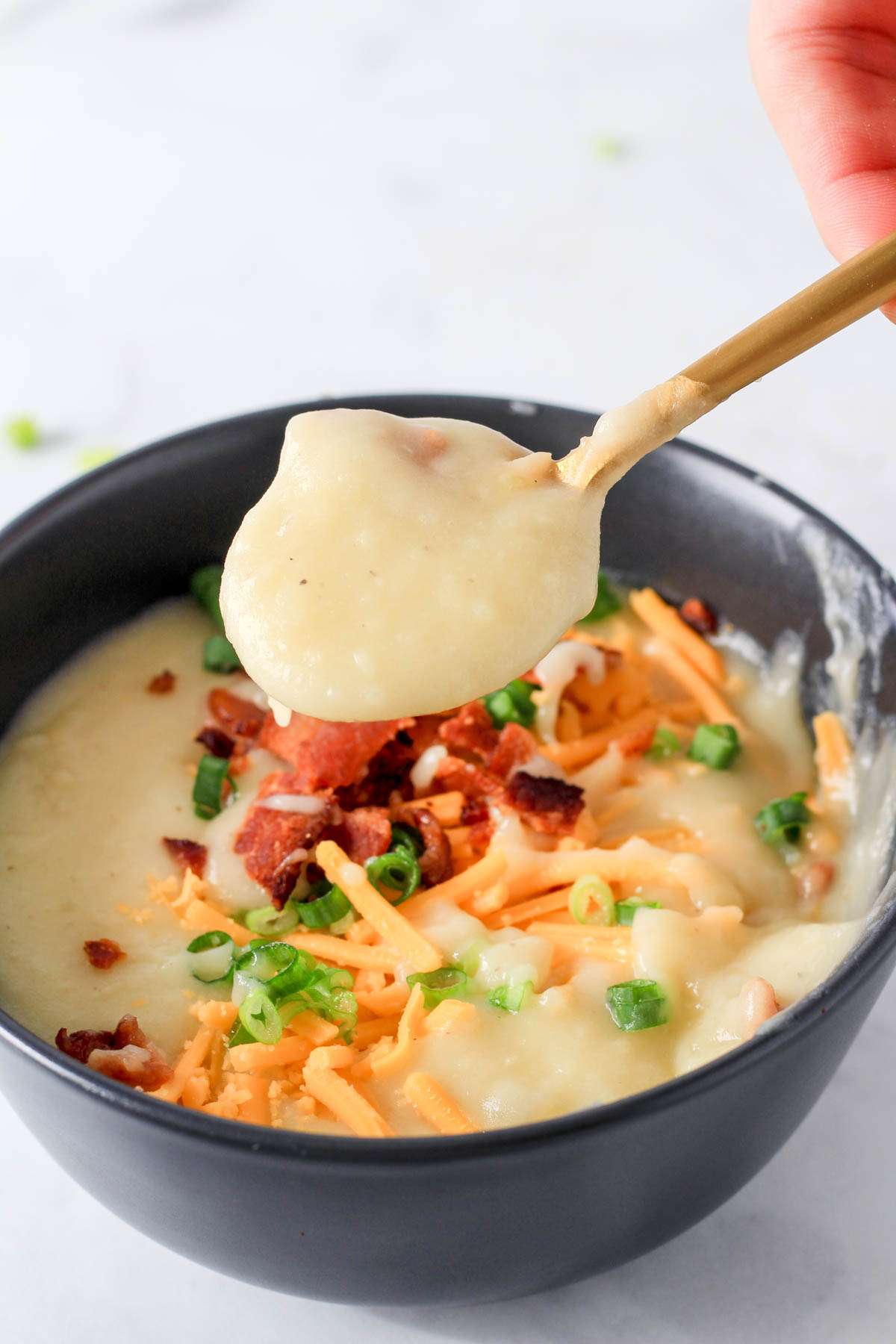 A gold spoon filled with creamy dairy-free potato soup in front of a blue bowl of soup topped with bacon, vegan cheese, and green onion.
