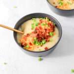A blue bowl filled with hearty and creamy dairy-free potato soup topped with vegan cheese, crispy bacon, and green onion with a gold spoon in the left side of the bowl.