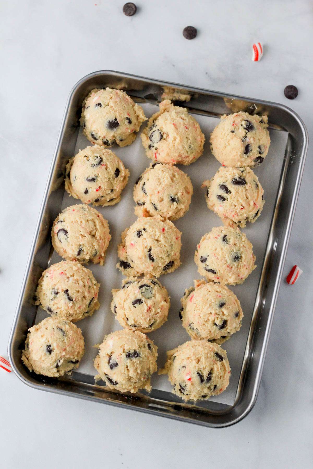 A silver rimmed baking pan lined with parchment paper and topped with balls of candy cane chocolate chip cookie dough.