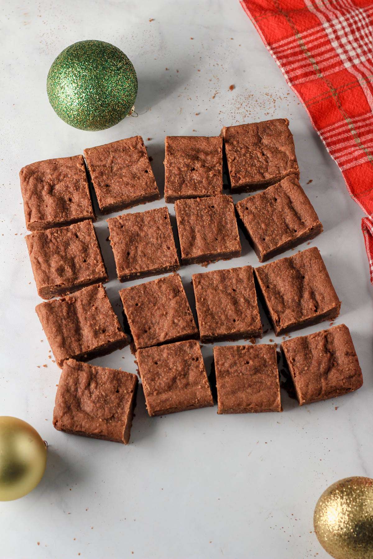 A white counter with 16 cut squares of vegan chocolate shortbread with three Christmas ornaments around the cookie bars.