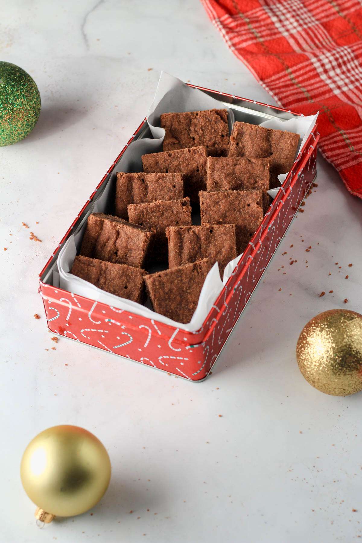 A red cookie box with parchment paper filled with two rows of chocolate shortbread bars on a white counter with a red towel.