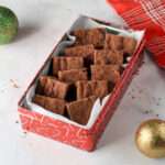 A red cookie tin with parchment paper filled with vegan chocolate shortbread squares on a white counter.