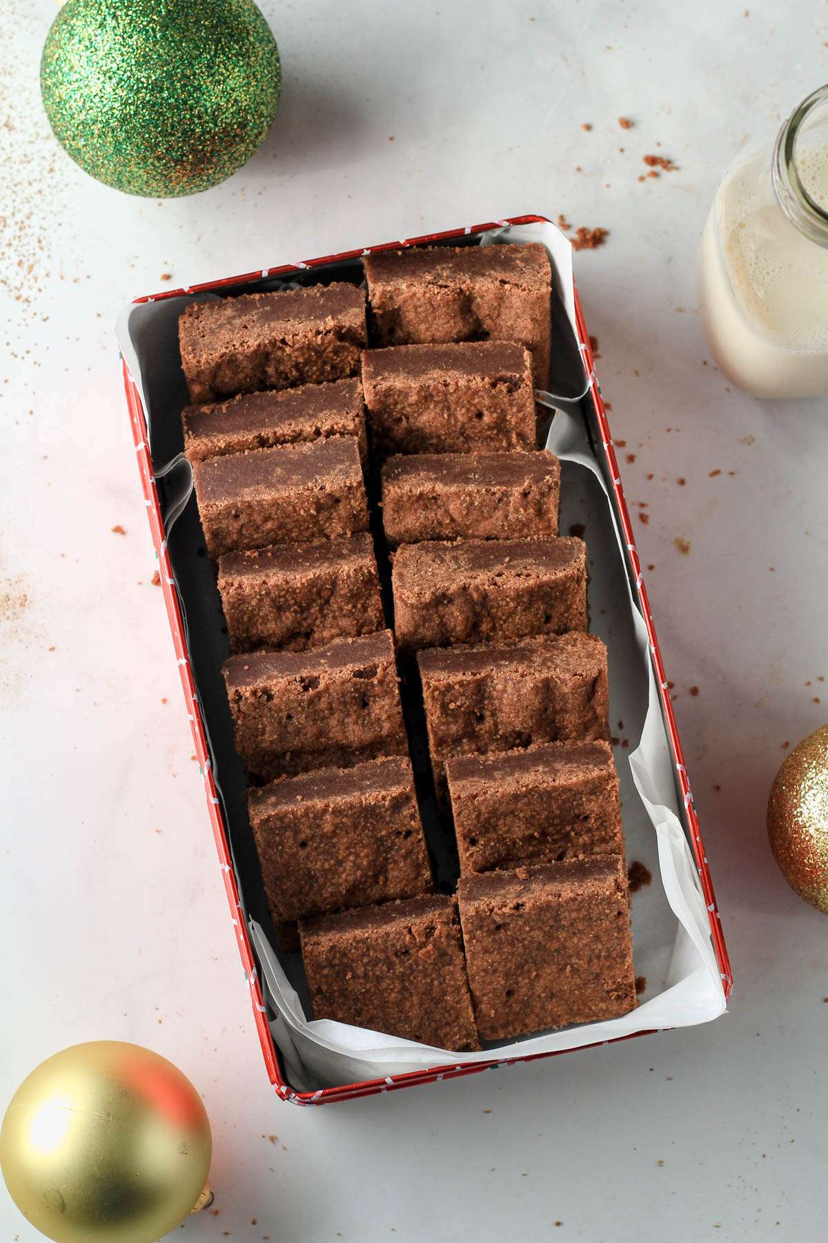 A red cookie box lined with parchment paper with two rows of chocolate shortbread cookies on a white counter.