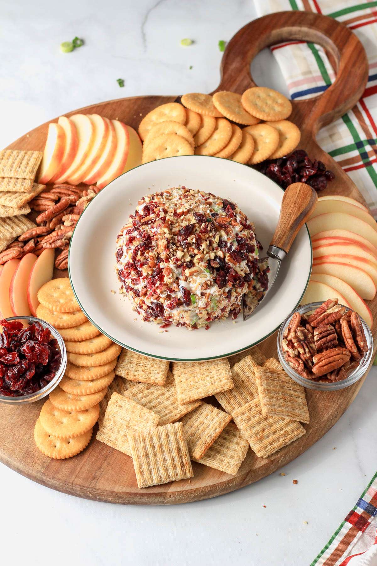A vegan cheese ball on a white plate in the middle of a wooden board topped with crackers, nuts, apples and more.